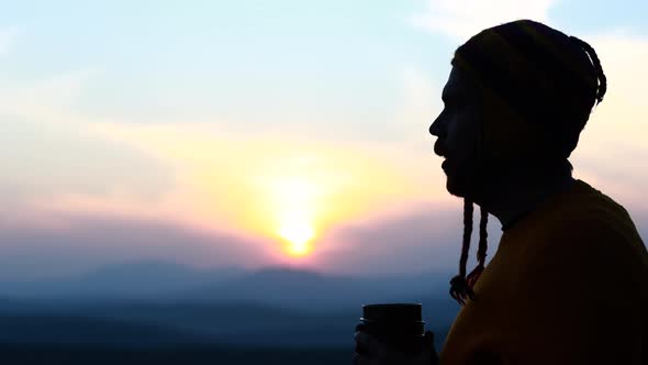 Man in Wooled Hat and Sweater Looking at Sun Dawn and Drinking Coffee in Nature