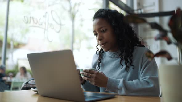 Pleased African woman wearing blue sweater working by laptop and drinking coffee