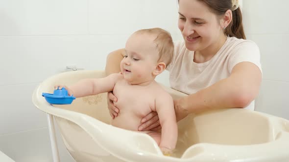 Smiling Mother Holding and Looking at Her Baby Son Playing in Small Children Bath at Home