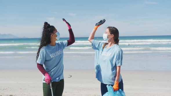 Two diverse women wearing volunteer t shirts and face masks picking up rubbish from beach