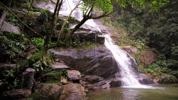 Tracking shot of a jungle cascading down a dark rocky outcropping into a dark green pool