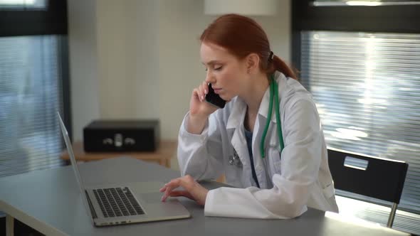 Side View of Serious Busy Female Doctor in White Coat Typing on Laptop and Talking on Mobile Phone