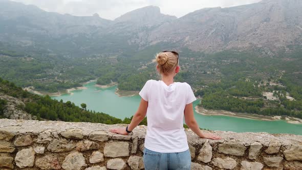 A Young Woman Looks at a Beautiful View with a Blue Lake and Mountains From the Wall of an Old