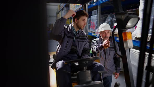 Young Handsome Caucasian Warehouse Loader Sitting in Forklift Discussing Plan with Curator