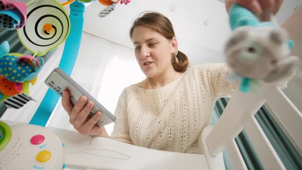 Busy Young Mother Shaking Colorful Rattle Toy to Her Baby While Using Smartphone