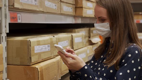 Woman in a Protective Mask Scans Boxes of Goods in a Warehouse with a Smartphone