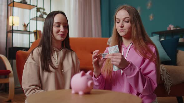 Girls Friends Siblings Sitting on Floor and Take Turns Dropping Dollar Banknote Into Piggy Bank