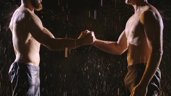 Two Muscular Topless Men Shake Hands in the Splashing Rain. Picture Taken in the Studio on a Black