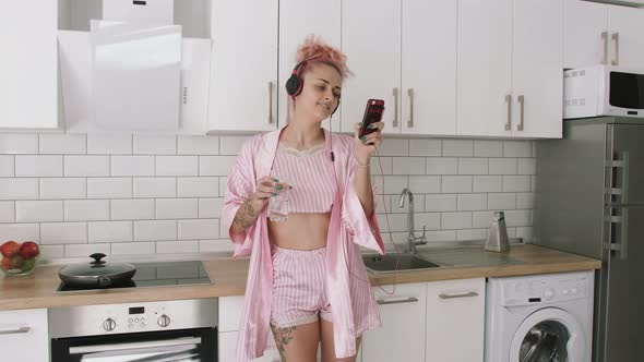 Happy Young Woman with Pink Hair Dancing in Kitchen Wearing Pink Pajamas and Listening to Music with