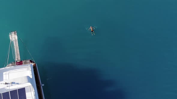 Aerial View of a Man Who Floats on His Back in the Turquoise Waters of the Sea Near a White Yacht