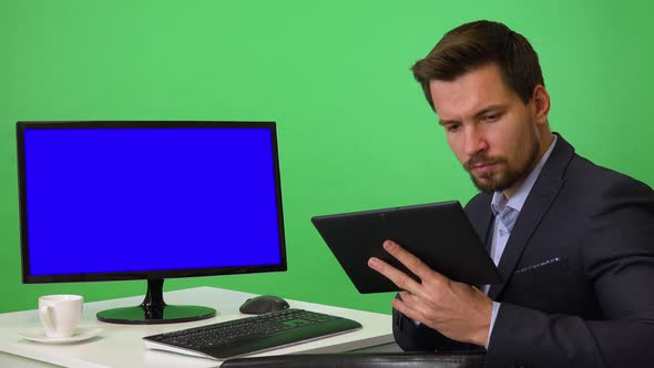 A Young Businessman Sits in Front of a Computer and Works on a Tablet - Green Screen Studio