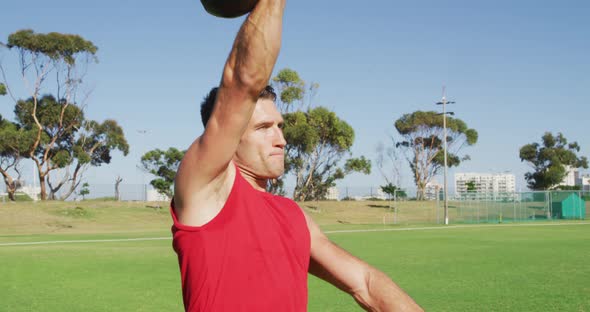 Fit caucasian man exercising outdoors, squatting and lifting kettlebell weight with one arm