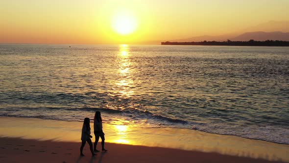 Young women walking on quiet exotic beach alongside sea reflecting beautiful sunlight of sunset with
