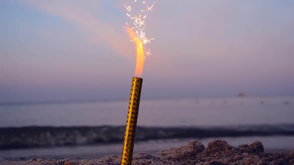 Burning Holiday Candle with Sparks on Beach in Sand on Background of Sea at Dusk