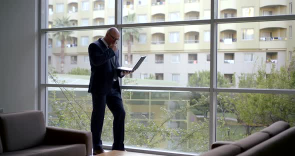 Bald Man Is Working with Notebook, Typing Messages, Standing Near Large Windows in Hall of Hotel