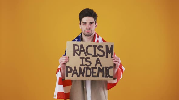 Man Wrapped in Flag of USA is Raising Up Cardboard Tablet with Inscription Racism is a Pandemic