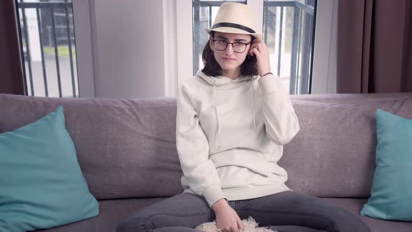 Girl in Glasses and a Hat Looks Straight Into the Camera Sitting Crosslegged on the Couch