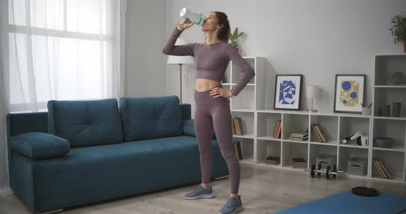 Sporty Woman Is Standing in Living Room and Relaxing Drinking Water Break at Morning Workout at Home