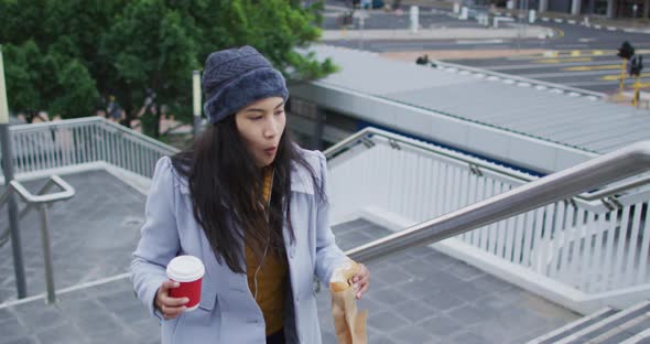 Asian woman walking holding takeaway coffee and eating sandwich