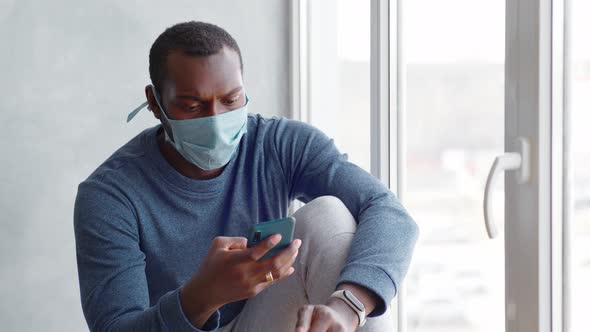 a Man with a Protective Mask on His Face During Quarantine Sits By the Window with a Smartphone