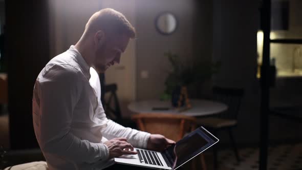 Confident Handsome Man Using Laptop While Sitting in Dark Living Room, Remote Working From Home.