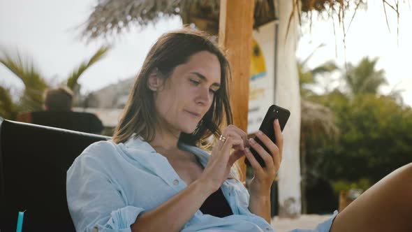 Happy Relaxed Tourist Woman Shopping Online Using Smartphone App Smiling, Resting in Exotic Sea