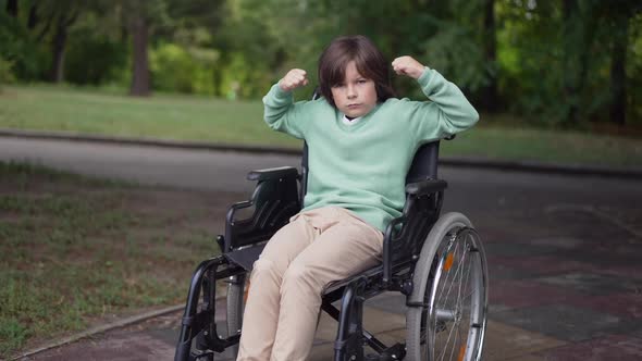 Zoom in Serious Caucasian Disabled Boy in Wheelchair Gesturing Strength Gesture Looking at Camera