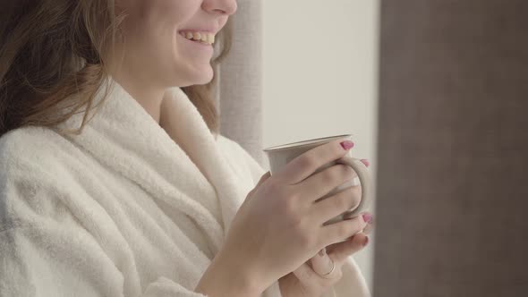 Close-up of Unrecognizable Young Caucasian Woman in White Bathrobe Holding Coffee Cup and Laughing