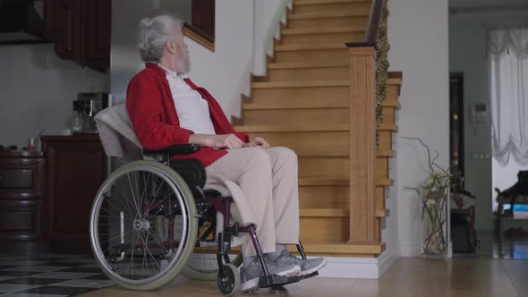 Sad Disabled Senior Man in Wheelchair Looking Up at Stairs in Nursing Home Looking at Camera with