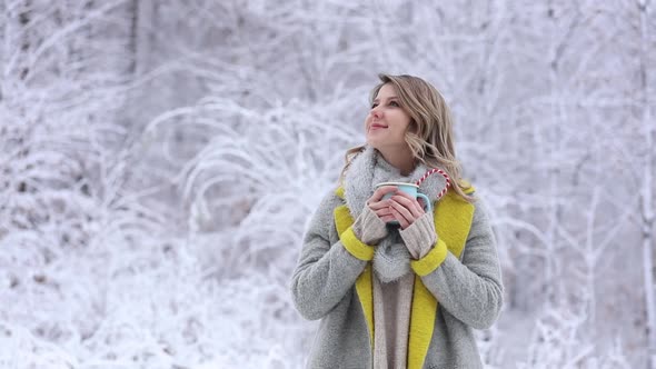 Beautiful woman in coat with cup of hot drink in a snow forest.