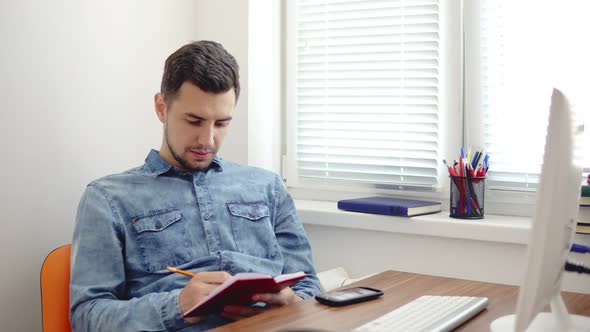 Young Businessman Sitting By the Computer in Stylish Modern Office and Taking Notes Using His Pencil