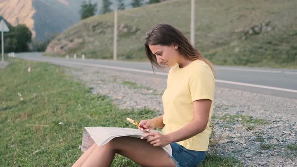 Beautiful Girl Tourist Looks at a Map of Roads While Sitting on a Highway in Summer Sunny Weather