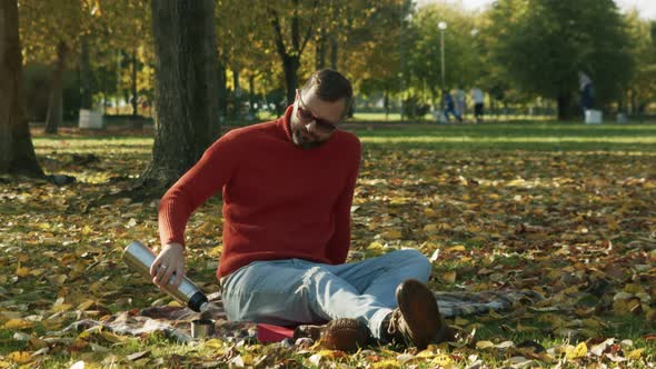 a Man in Black Glasses and an Orange Sweater in a Public Park Sits on the Autumn Grass Pouring