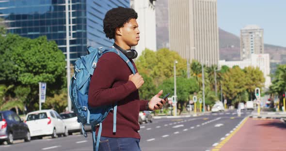 African american man in city, using smartphone, wearing headphones and backpack crossing street