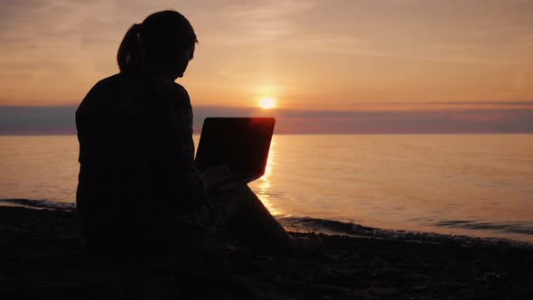 Silhouette of a Woman Working with a Laptop By the Sea at Sunset