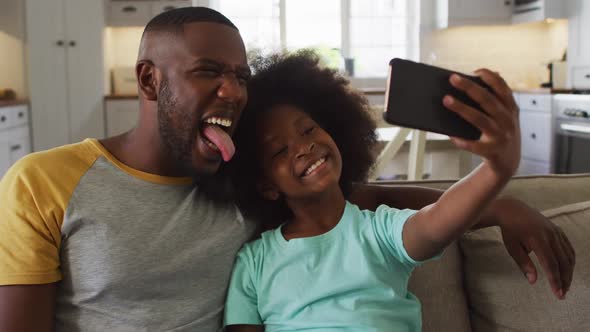 African american daughter and her father making faces taking selfie together on couch