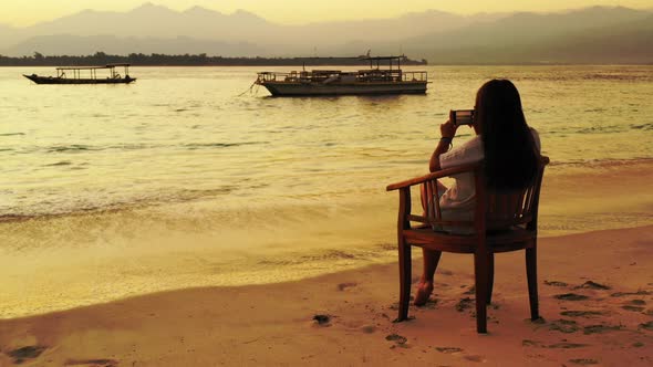Young woman sitting on exotic beach taking pictures of beautiful calm lagoon with anchored boats at