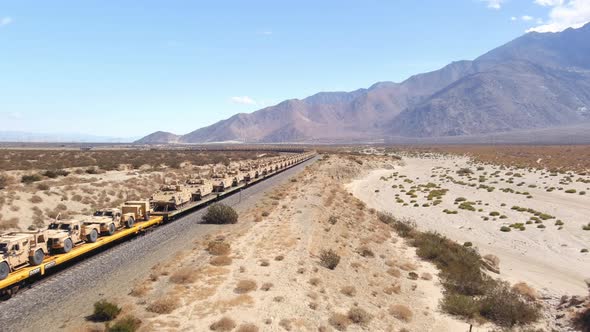 Tracking aerial shot of train carrying US Army warfare vehicles through the desert landscape in Cali