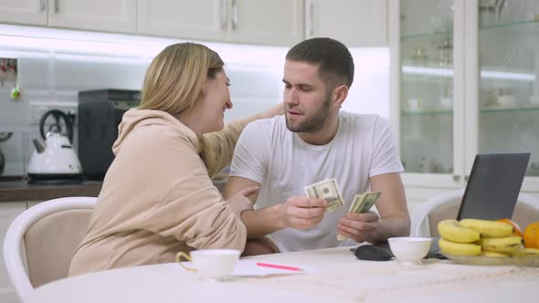 Portrait of Young Caucasian Husband Giving Money to Smiling Wife Sitting in Kitchen at Home