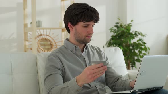 Young Man is Sitting on the Sofa in the Living Room Making Online Purchases Through a Laptop Holding