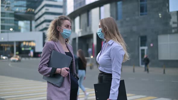 Two women in medical masks are talking on the street next to an office building