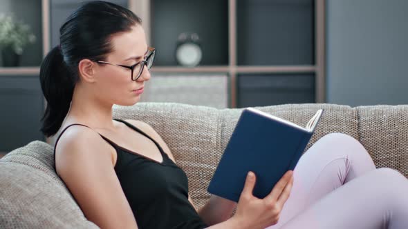 Confident Young Asian Woman Reading Interesting Book Relaxing at Home Lying on Comfortable Couch