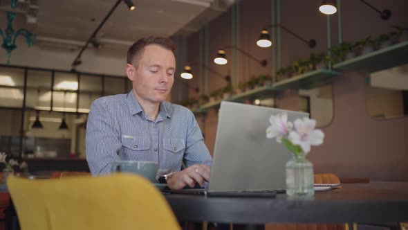 A Young Man in a Shirt is Sitting at a Table with a Laptop and Typing on the Keyboard