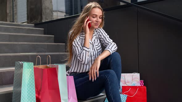 Girl Sitting on Stairs with Bags Talking on Mobile Phone About Sale in Shopping Mall in Black Friday