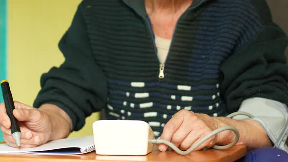 Caucasian Pensioner sitting at the table at home measures blood pressure and writes down the reading