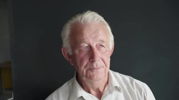 Portrait of Senior Man Looks at Camera Thoughtfully on Background