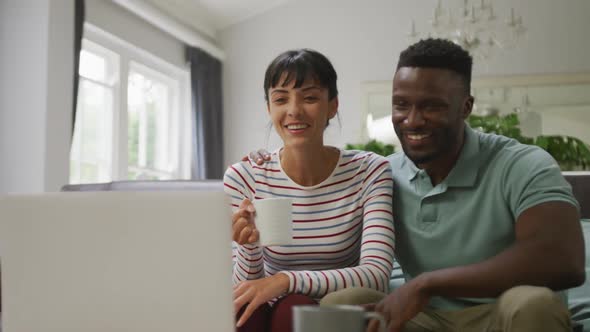 Happy diverse couple sitting on couch and using laptop in living room