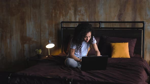Young Attractive AfricanAmerican Woman Using Laptop Computer in the Bed in Her Dark Loft Bedroom