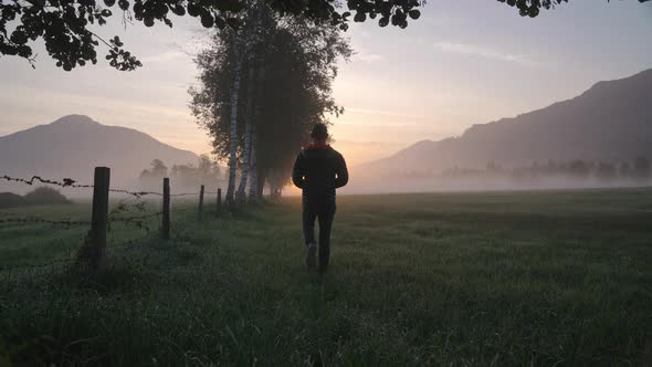 Man Walking Through Meadow Towards Foggy Landscape And Sunrise