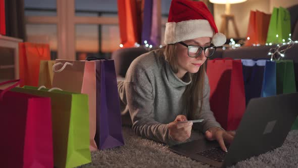Happy Woman with Glasses Wearing a Santa Claus Hat Is Lying on the Carpet and Makes an Online